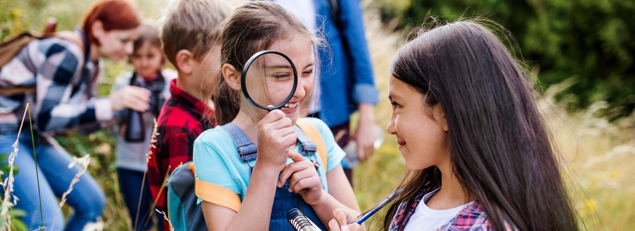 Girls outdoors exploring; one holding a magnifying glass and the other looking at the girl with a notepad